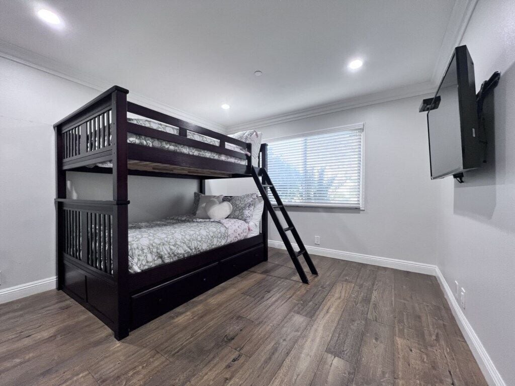 A bunk bed with a ladder, a window with blinds, and a flat-screen TV mounted on the wall.