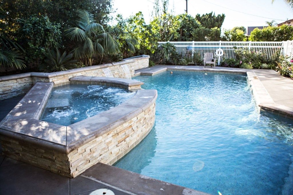 A swimming pool with a hot tub, surrounded by a white fence and lush greenery.