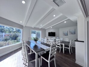 A dining area with a long table, white chairs, a flat-screen TV, and a view of the outdoors.