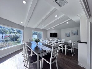 Comedor con una mesa larga, sillas blancas, televisor de pantalla plana y vista al exterior.