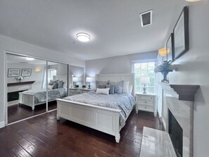 A bedroom with a large bed, a fireplace, a mirror, and a window.