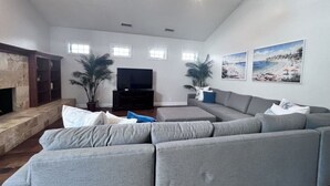 A spacious living room with a large gray sectional sofa, a flat-screen TV, a stone fireplace, and two potted plants. - Fantastic 5 Bedroom 4 Bath Home With new Pool & Jacuzzi (Anaheim)