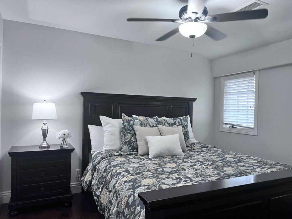 Une chambre à coucher comprenant un lit, une table de chevet, une lampe, un ventilateur de plafond et une fenêtre avec des stores.