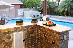 A poolside bar with a stone countertop, a small refrigerator, and a decorative flower arrangement. - Amazing Large Family Home W/pool, 1 Mile to Disney (Anaheim)