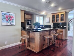 A kitchen with wooden cabinets, a central island with stools, a large window, and a staircase in the background. - Amazing Large Family Home W/pool, 1 Mile to Disney (Anaheim)