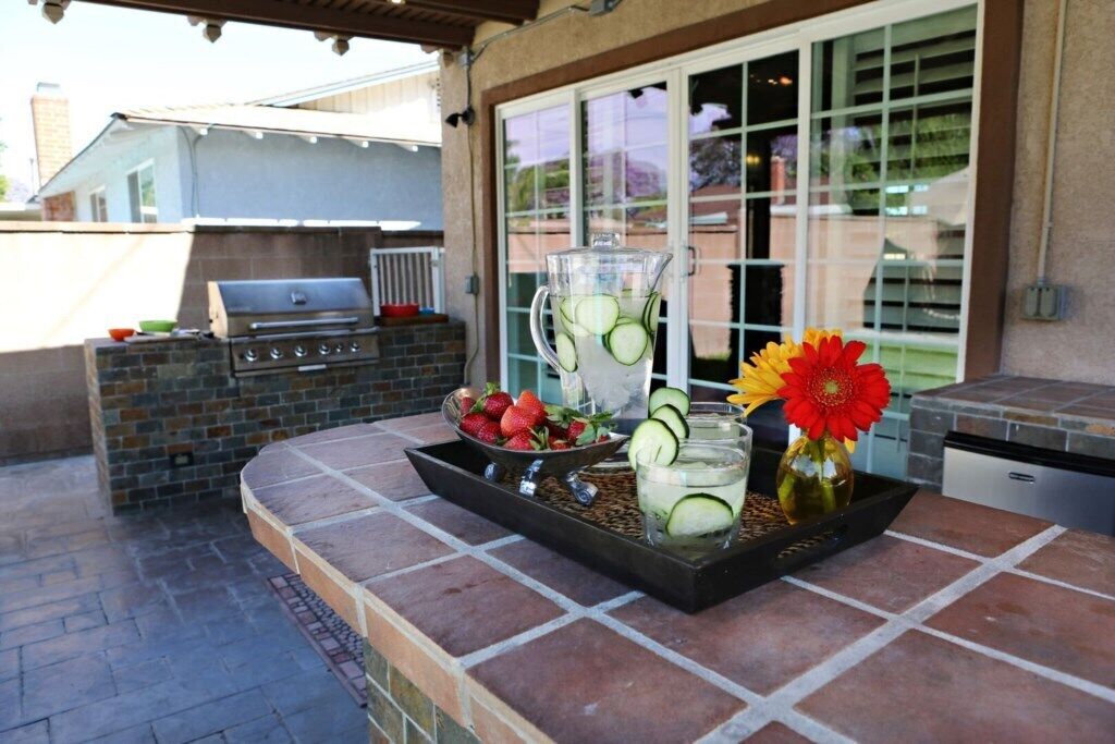 Een terras buiten met een barbecue, een dienblad met fruit en een kan, en een vaas met bloemen.