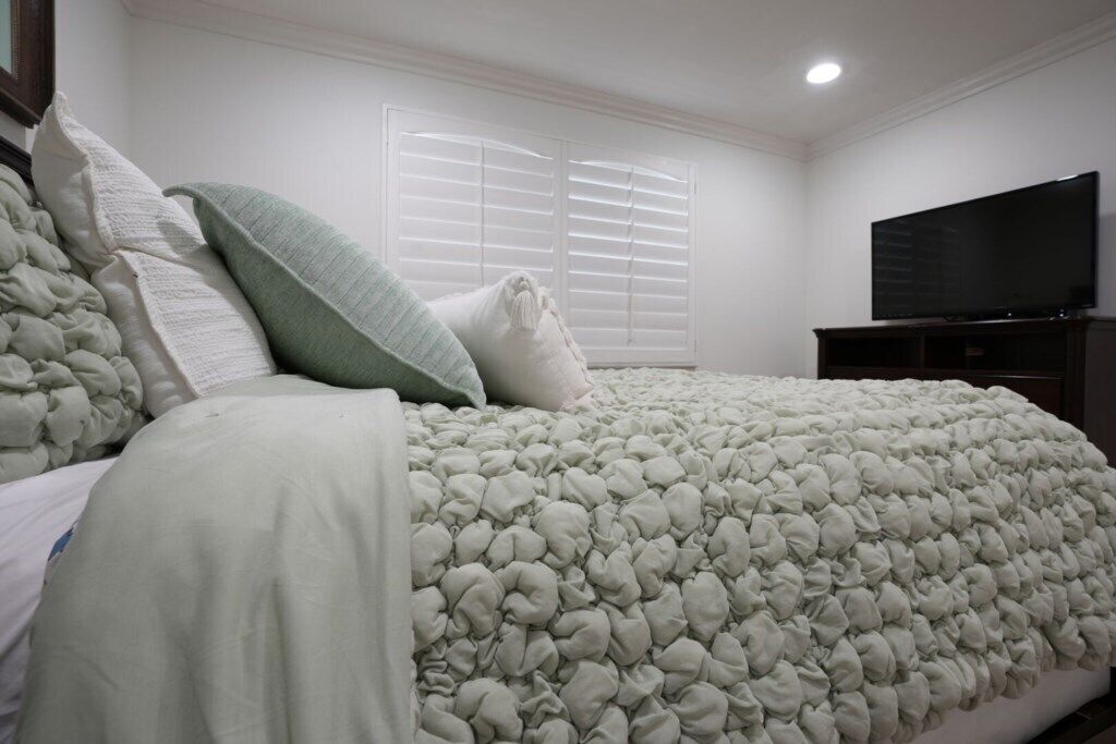 A bedroom with a bed, pillows, a television, and a window with blinds.