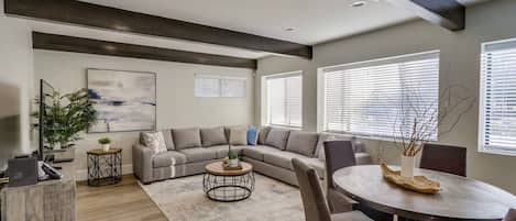 A modern living room with a sectional sofa, a round coffee table, a dining table with chairs, and a large window with blinds.