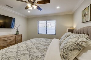 Un dormitorio con cama, ventilador de techo, ventana con persianas, televisor montado en la pared y cómoda de madera.