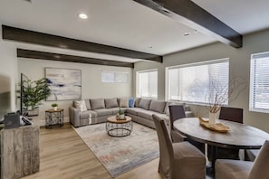A modern living room with a sectional sofa, a round coffee table, a dining table with chairs, and a large window with blinds. - 5 Bedroom plus loft room, 3 Bath home (Fullerton)