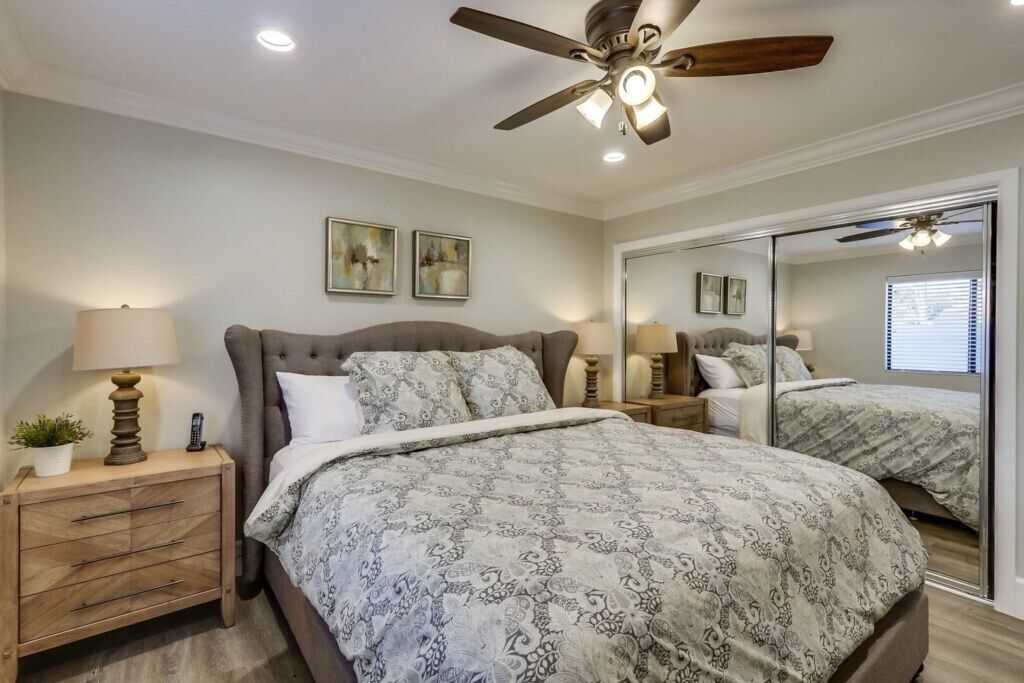 Un dormitorio con cama, mesitas de noche, ventilador de techo y un espejo grande.