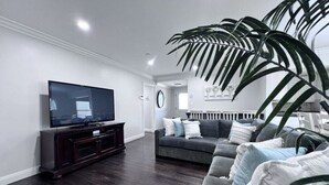 A living room with a dark wood floor, a large flat-screen TV on a dark wooden stand, a grey sofa with multiple cushions, and a large green plant. - Dazzling 5 Bedroom Pool Home 1/4 Mile To Disney (Anaheim)