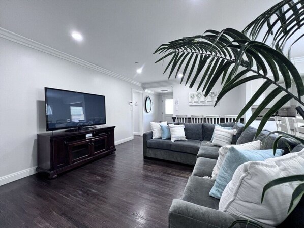 A living room with a dark wood floor, a large flat-screen TV on a dark wooden stand, a grey sofa with multiple cushions, and a large green plant.