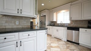 A kitchen with white cabinets, a stainless steel refrigerator, a dishwasher, and a window with blinds. - Dazzling 5 Bedroom Pool Home 1/4 Mile To Disney (Anaheim)