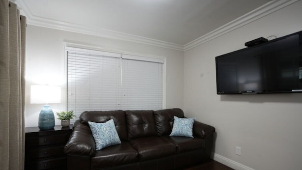 A living room with a brown leather sofa, two patterned cushions, a small table with a vase and plant, a television on the wall, and a window with blinds. - Awesome Pool Home 1/4 Mile To Disney Tram on Magic Way (Anaheim)