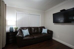 A living room with a brown leather sofa, two patterned cushions, a small table with a vase and plant, a television on the wall, and a window with blinds. - Awesome Pool Home 1/4 Mile To Disney Tram on Magic Way (Anaheim)