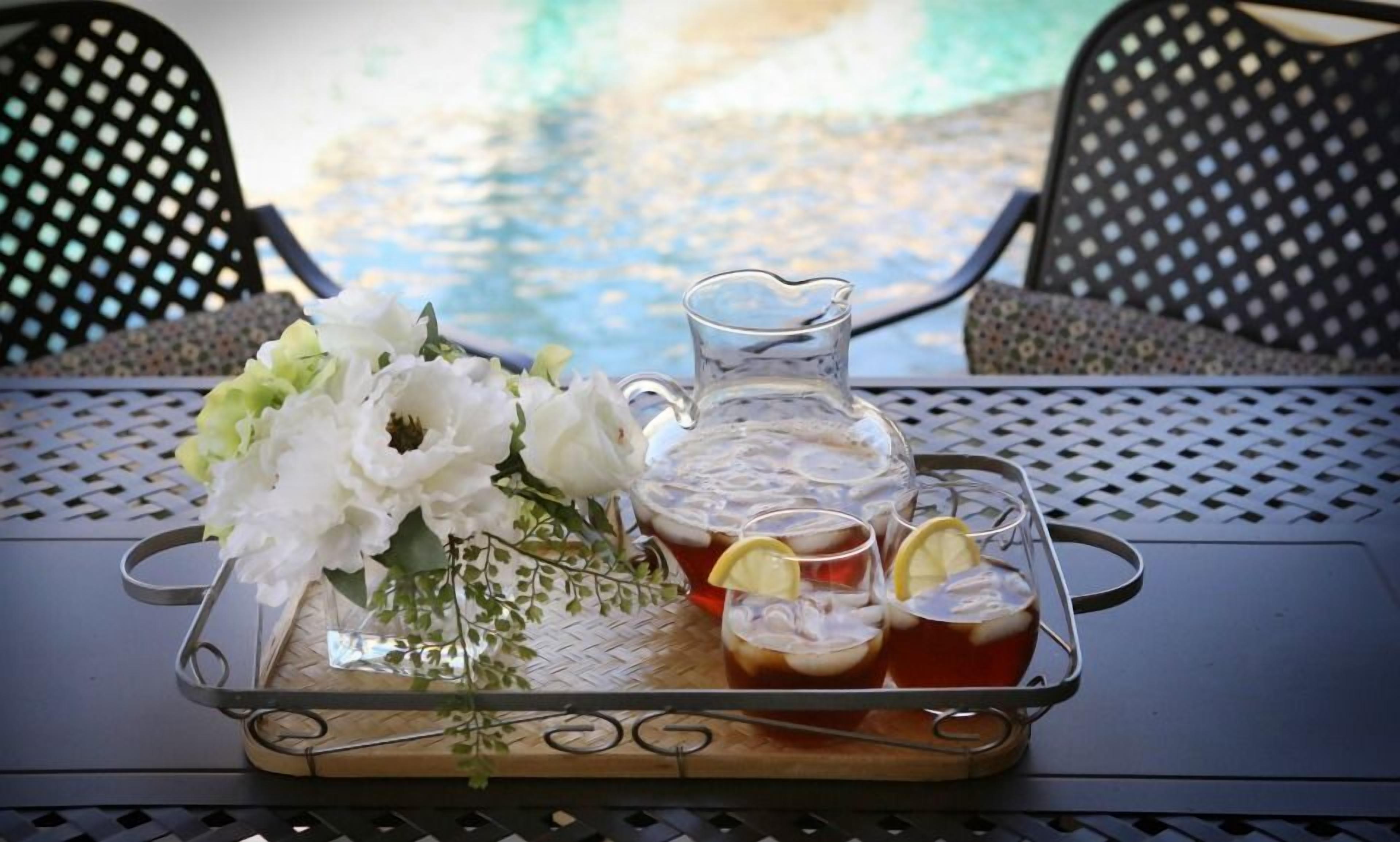 Un plateau avec une carafe de thé glacé, des verres et un bouquet de fleurs blanches, le tout posé sur une table d’où l’on a vue sur une piscine.