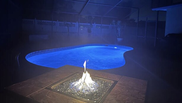 Une piscine avec un foyer au centre, entourée d’une terrasse et d’une clôture blanche en arrière-plan.