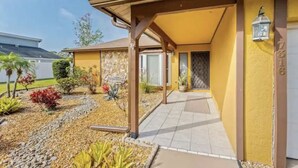 Un bâtiment jaune doté d’une entrée abritée, d’un mur en pierre et d’un petit jardin où poussent différentes espèces de plantes.