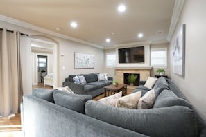 A living room with a grey sofa set, a flat-screen TV mounted above a fireplace, and a painting on the wall.