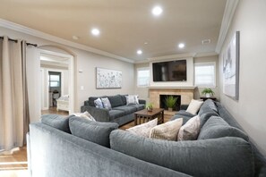 A living room with a grey sofa set, a flat-screen TV mounted above a fireplace, and a painting on the wall.