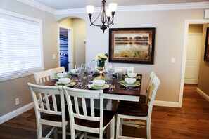 A dining room with a wooden table set for six, white chairs, a chandelier, and a framed painting on the wall.
