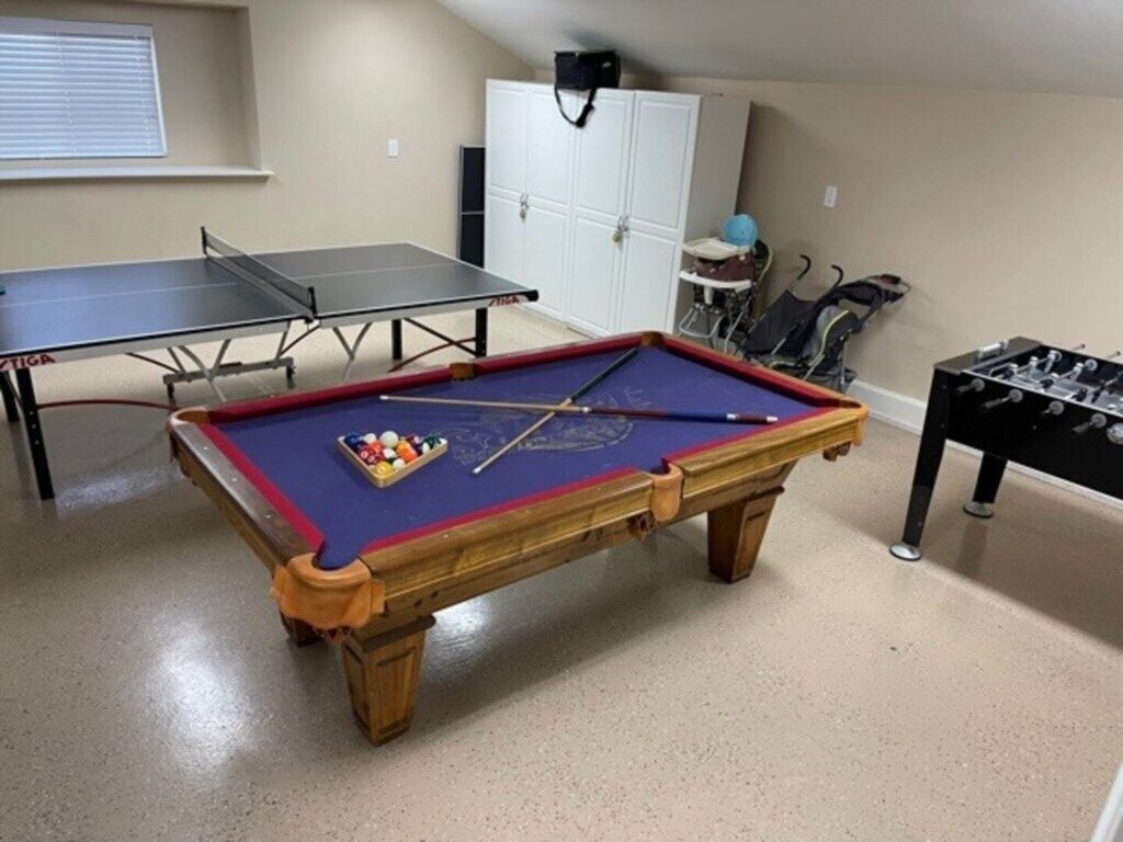 Ett biljardbord med bollar och biljardköer, ett bord för bordtennis och ett fotbollsspel i ett rum med ett fönster och ett vitt skåp.