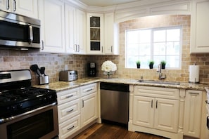 A kitchen with white cabinets, a stainless steel stove, a dishwasher, a window with plants, and a brick backsplash.