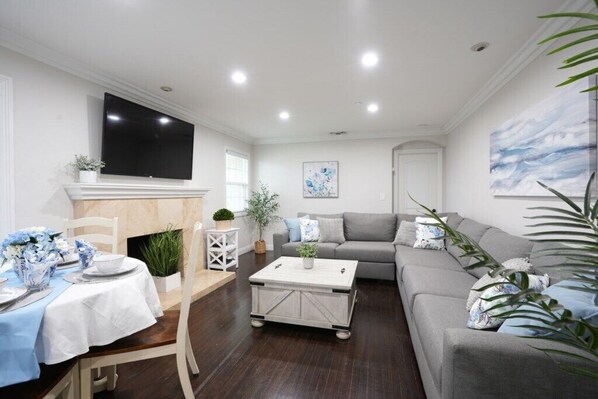 A living room with a sofa, a coffee table, a TV mounted on the wall, and a dining area with a table set for two. - Incredible 5 Bedroom, 4 Bath, Rock Pool & Jacuzzi Home, 1/2 Mile Walk To Disney (Anaheim)