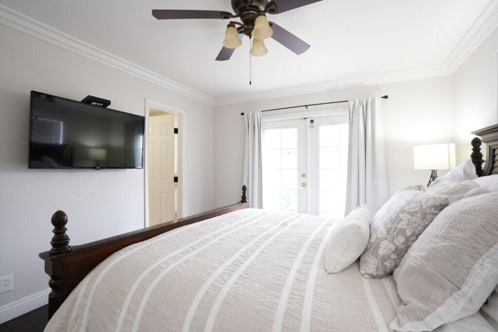 Ein Schlafzimmer mit einem Bett, einem Deckenventilator, einem an der Wand befestigten Fernseher und einer Tür zu einem anderen Raum.
