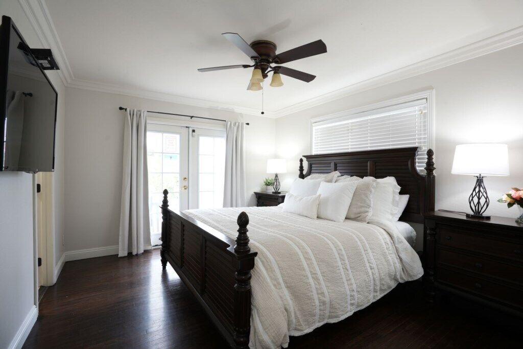 A bedroom with a dark wooden bed, a ceiling fan, a window with blinds, a nightstand with a lamp, and a dresser.