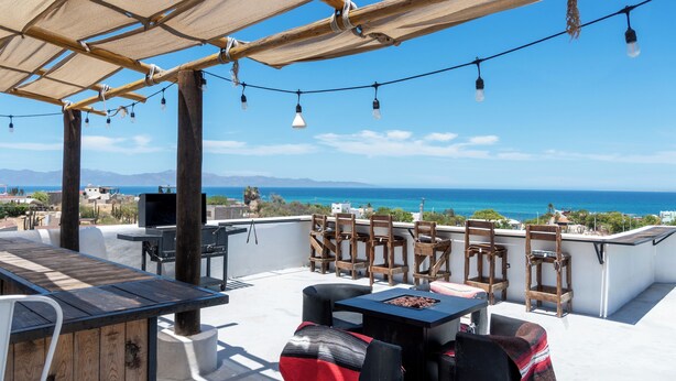 An outdoor patio with a wooden table and chairs, a grill, and a view of the ocean. - Casa Arrecife| Tortuga: Wifi+ Cozy, Private bathroom, 1 room (La Ventana)