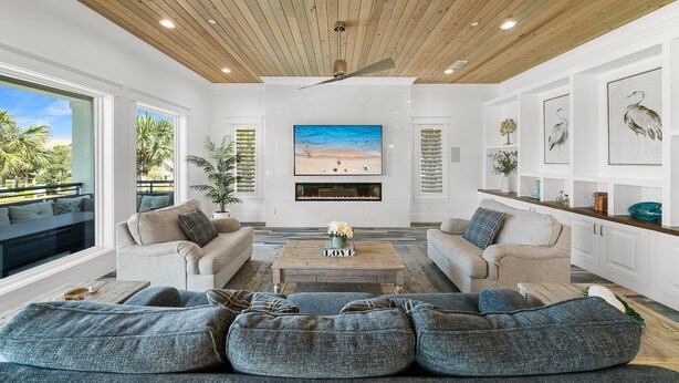 Un salón amplio con techo de madera, ventanas grandes, chimenea y vistas a palmeras.