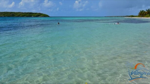 Een strand met helder turquoise water, een zandstrand en een eiland in de verte.