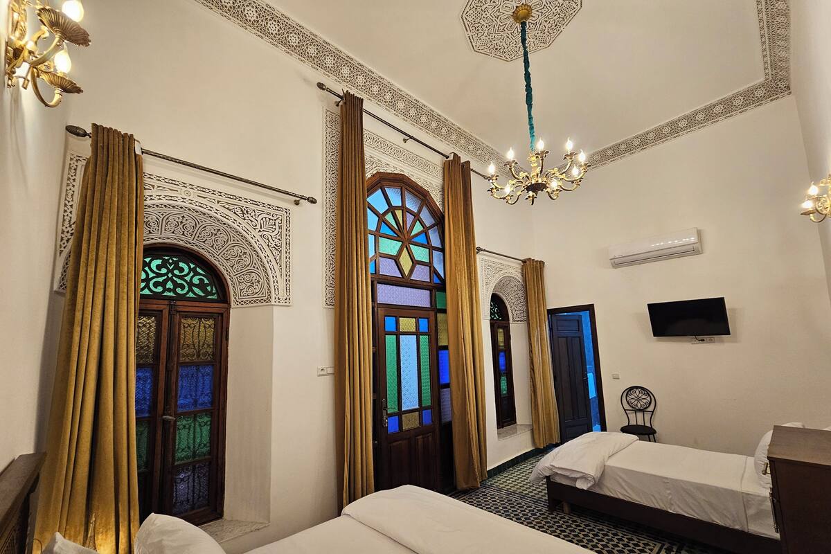 A hotel room with two beds, a chandelier, a TV, and decorative windows.