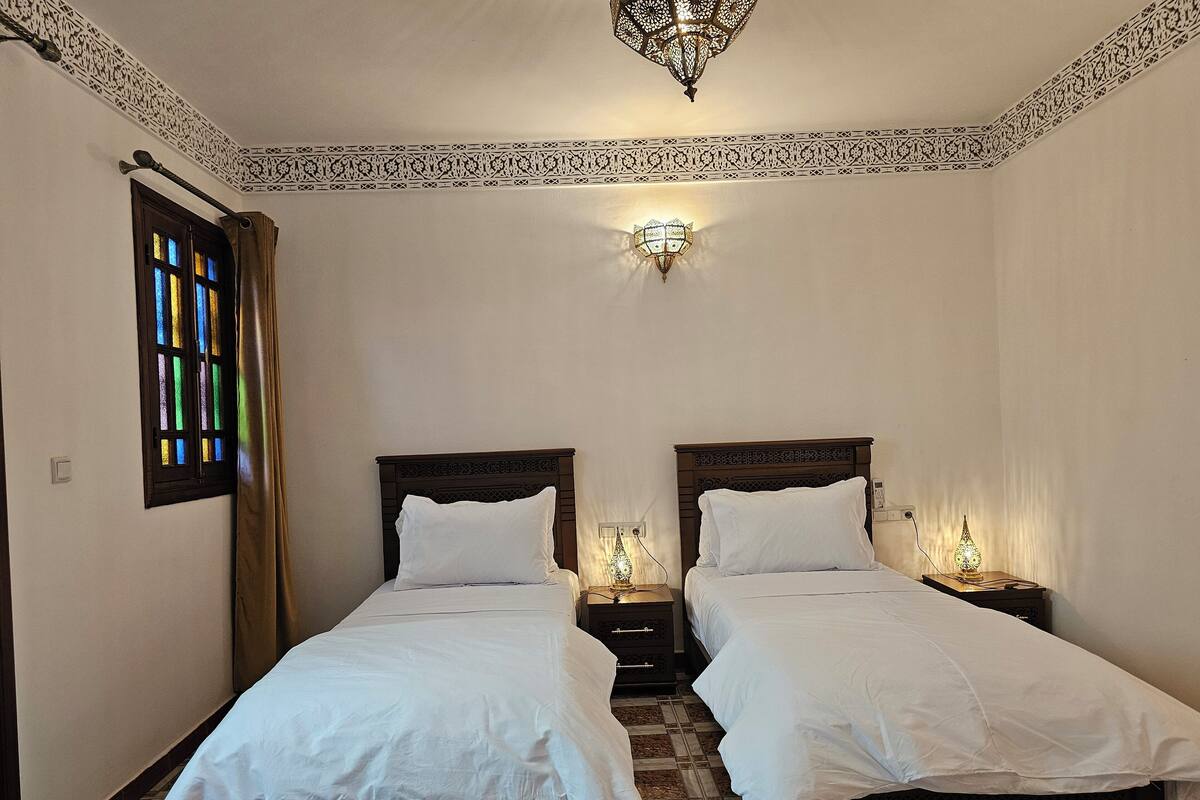 A room with two beds, a decorative ceiling, a wall-mounted light fixture, and a window with stained glass.