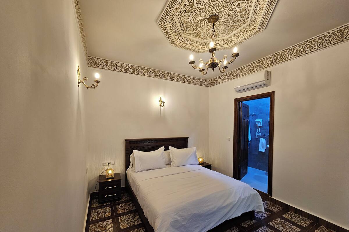 A bedroom with a large bed, a nightstand, a wall-mounted lamp, and a patterned floor.