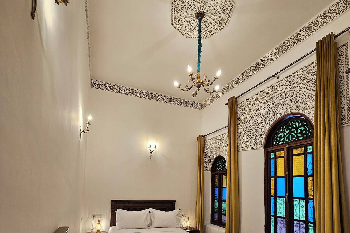 A hotel room with two beds, decorative wall sconces, a chandelier, and a stained glass window.