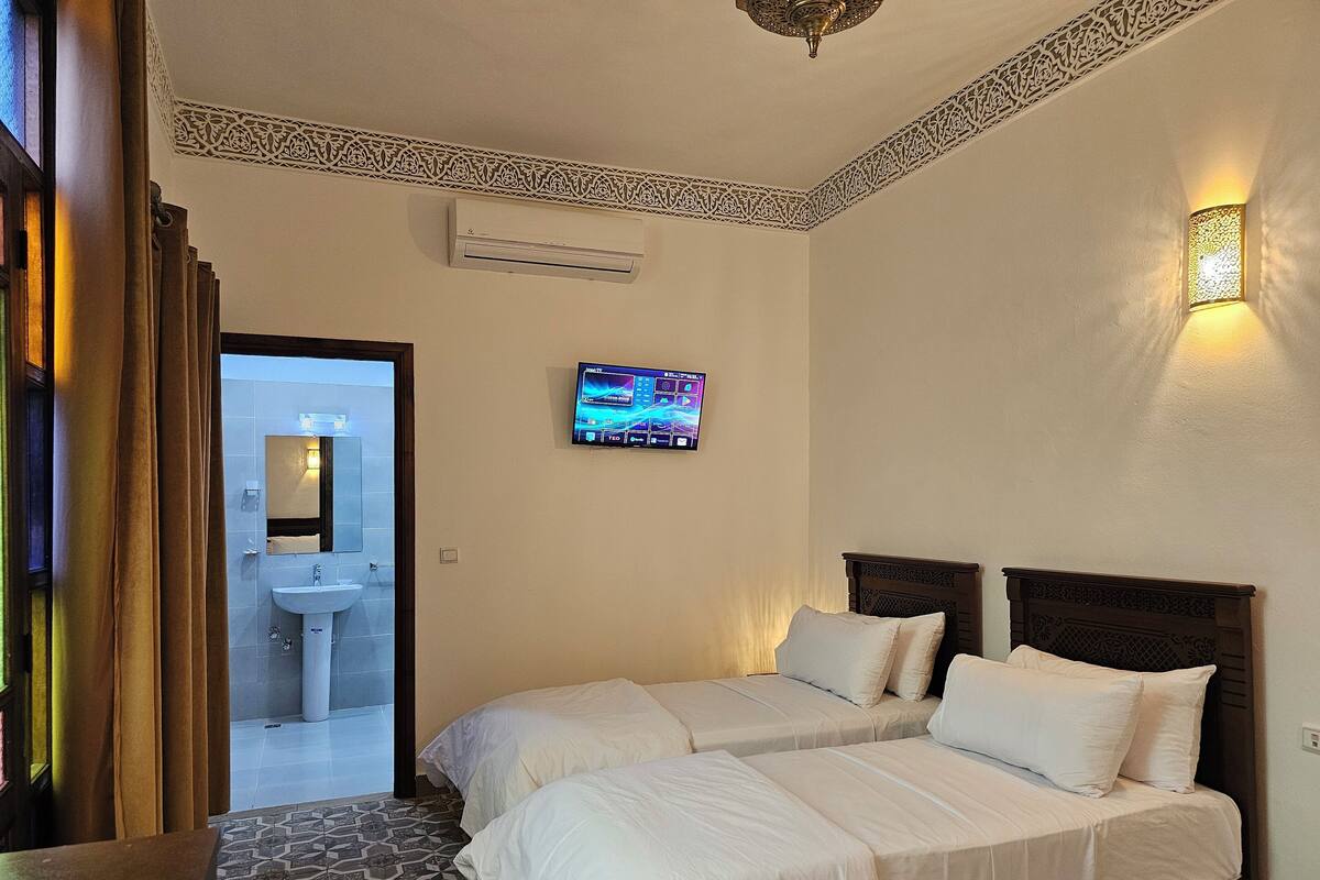 A hotel room with two beds, a TV, an air conditioning unit, and a bathroom visible through an open door.