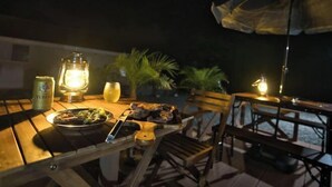 An outdoor dining area with a table set for two, featuring a plate of food, a drink, and a can of beer. There are two lantern-style lights providing illumination. - Racine Yakushima (Yakushima)