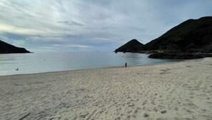 A beach with a person walking, calm water, and rocky hills in the background. - Racine Yakushima (Yakushima)