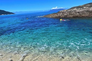 Una masa de agua costera cristalina, con terreno rocoso y montañas a lo lejos.
