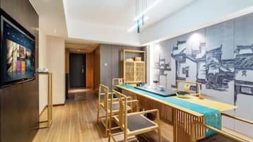 Habitación de hotel moderna con piso de madera, zona de comedor con una mesa larga y sillas, televisor de pantalla plana y pared con un mural de un paisaje urbano.