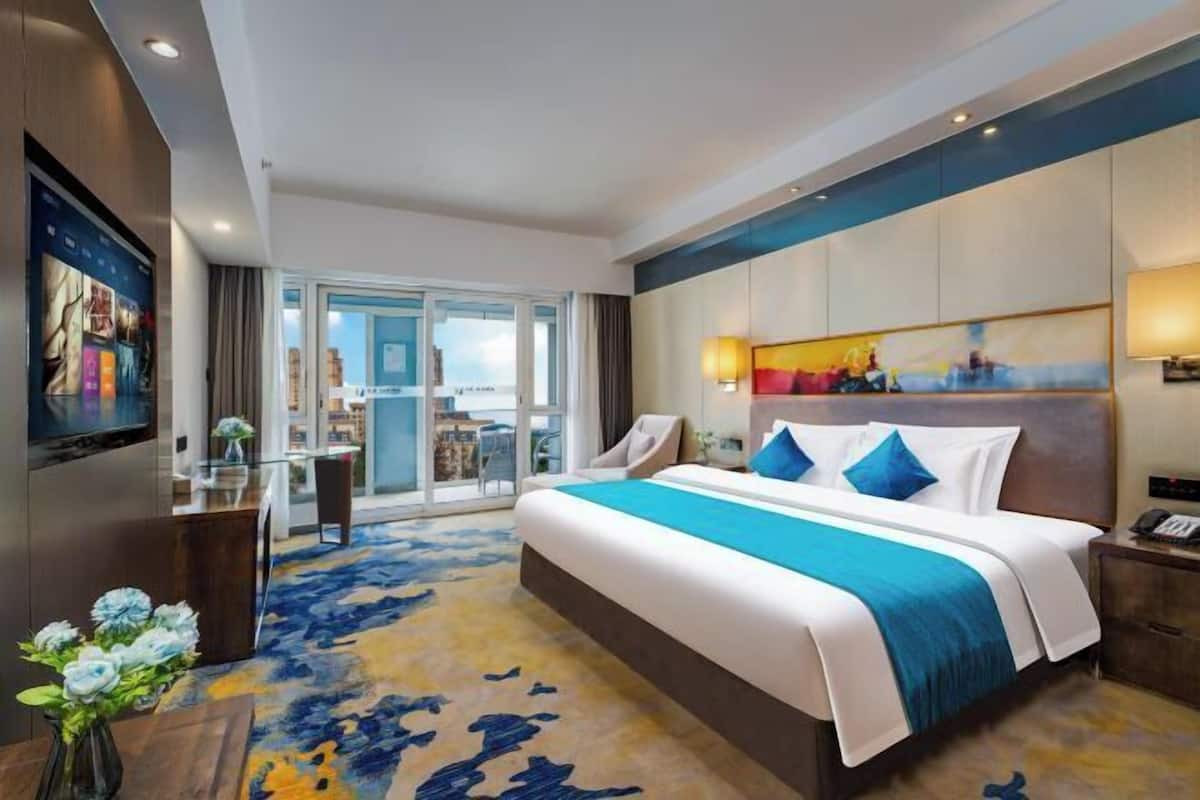 A hotel room with a large bed, a desk with a chair, a TV, and a view of the city through large windows.