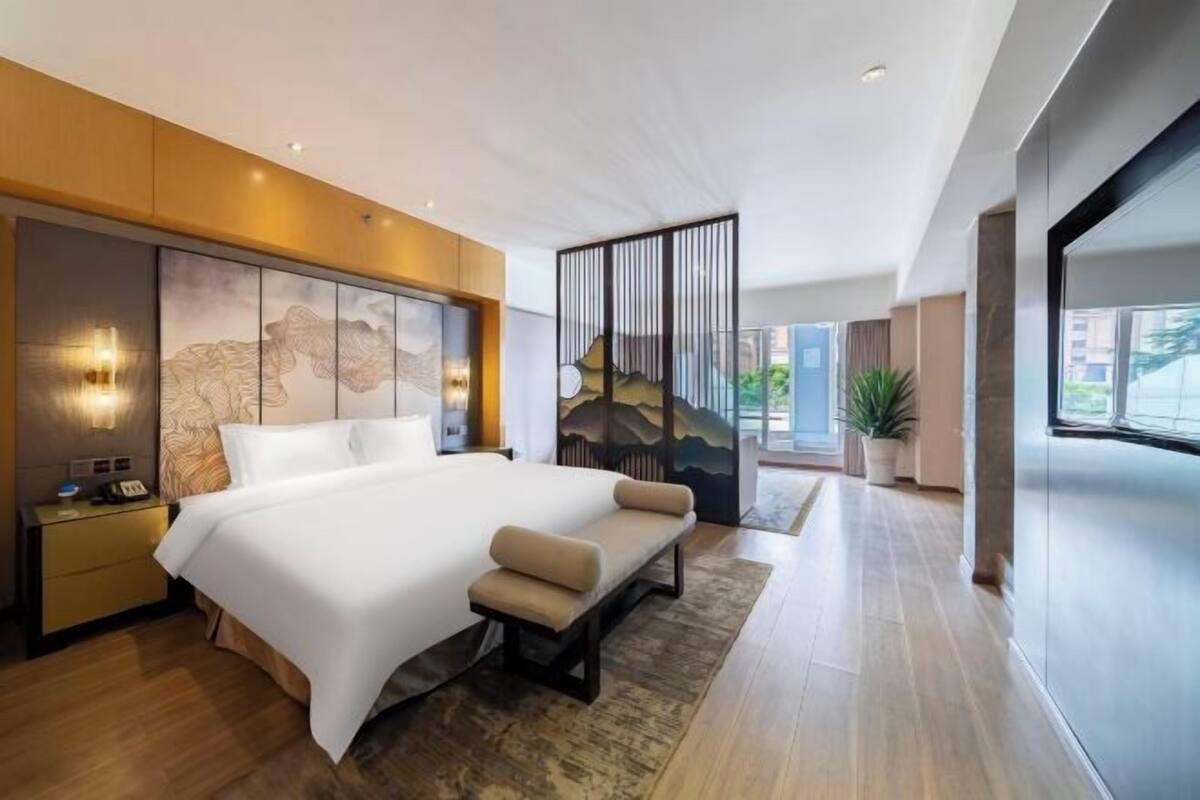A modern hotel room with a large bed, bedside tables, a bench, a decorative wall, and a large window.