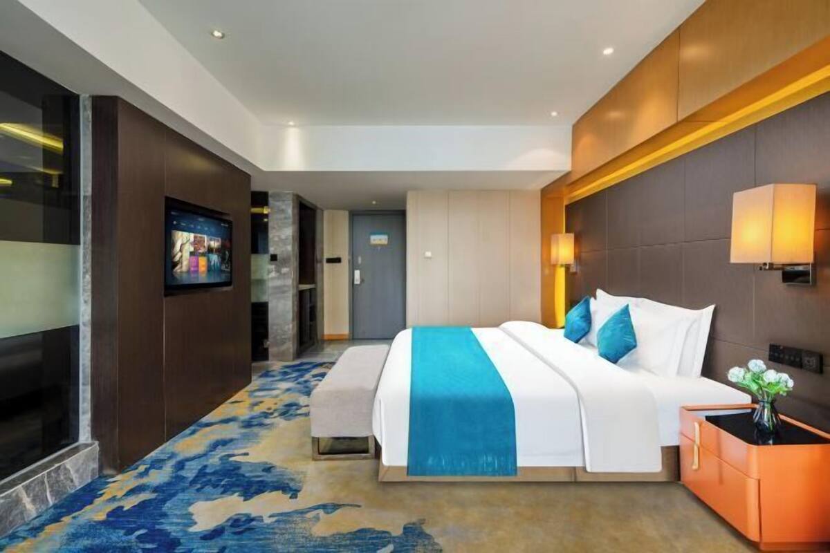 A modern hotel room with a large bed, a television, a desk with a lamp, and a carpeted floor.