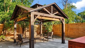 Una pérgola de madera con techo, zona de estar con sillas y una mesa, y un banco rojo.