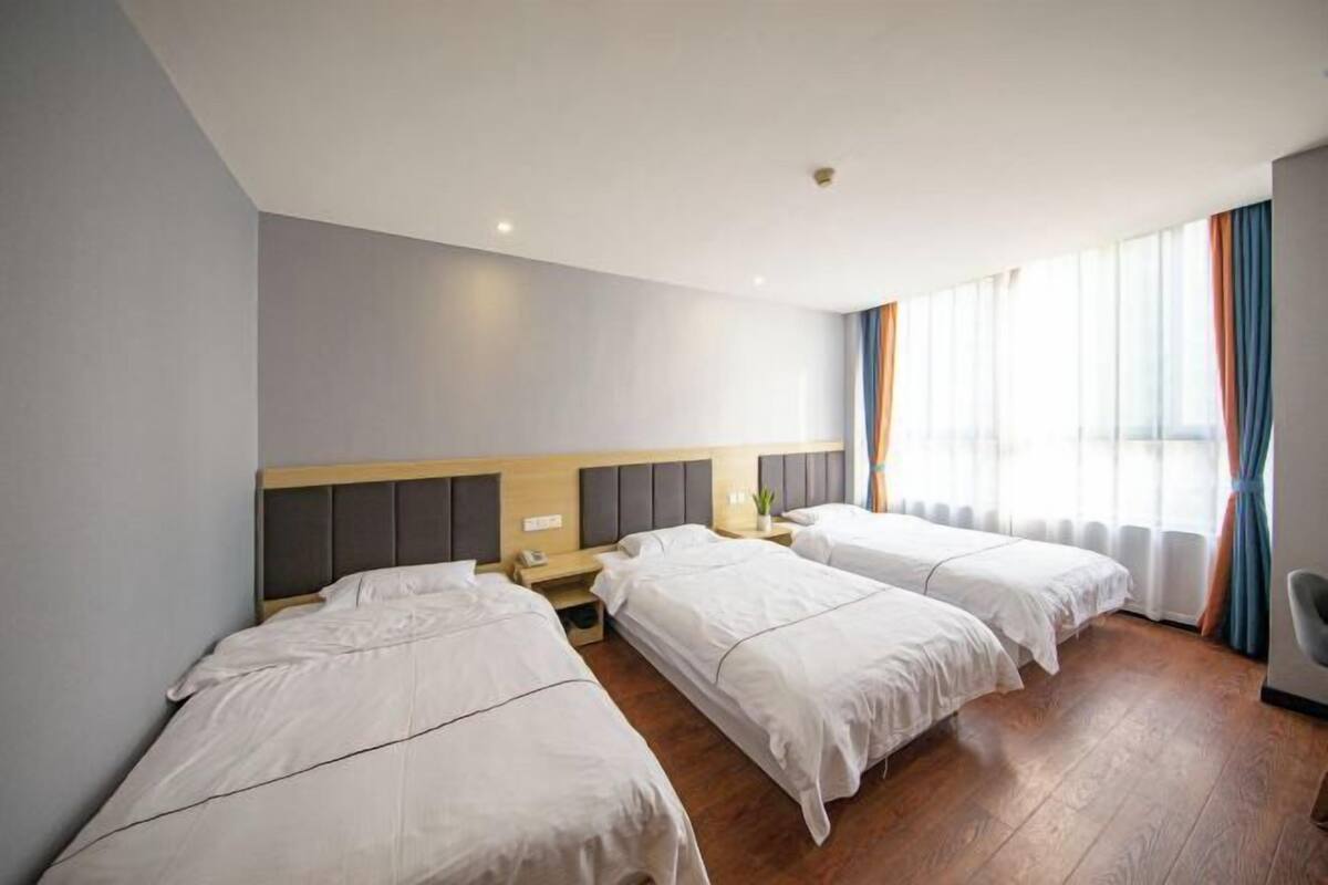 A hotel room with three single beds, a wooden headboard, a bedside table with a lamp, and a large window with curtains.