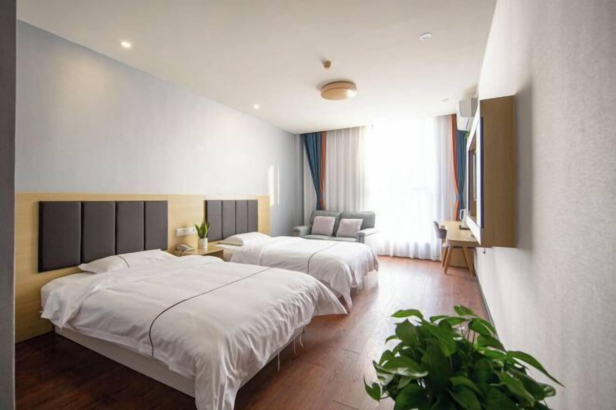 A modern hotel room with two beds, a desk, a chair, a TV, and a large window with curtains.