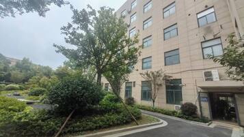 A multi-story building with a paved entrance area, surrounded by greenery and trees.
